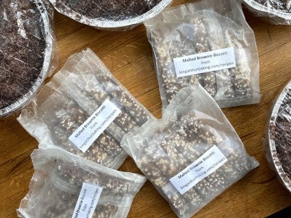 Malted Brownie Biscotti bagged in waxed paper bags and labeled, on a wooden countertop.